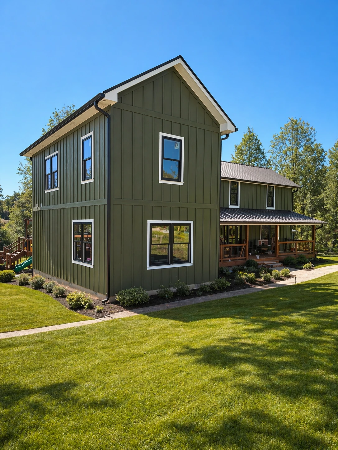 Homestead Place farmhouse front corner view — dark green board-and-batten with wraparound covered porch and metal roof